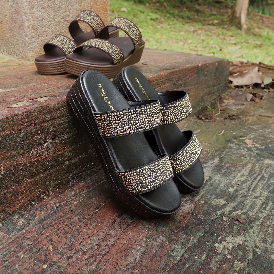 Thomas Chan Strappy Rhinestone Flatform Sandals in black, featuring dual straps embellished with shimmering rhinestones. The sandals rest against a rustic outdoor surface, with another pair of the same design in brown visible in the background. The shoes have a cushioned flatform sole, designed for comfort and style