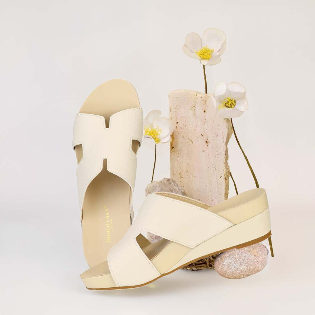 The image shows a pair of stylish Thomas Chan ivory white color comfortable Pebble Lychee H-Strap Low Wedge Slides sandals. These shoes feature a simple yet casual H-strap design, providing both elegance and comfort. The wedges are from Thomas Chan Malaysia, as indicated by the branding inside the shoe. The setting is enhanced by the presence of a few blooming flowers and a stone prop in the light background, giving the image a minimal and artistic feel