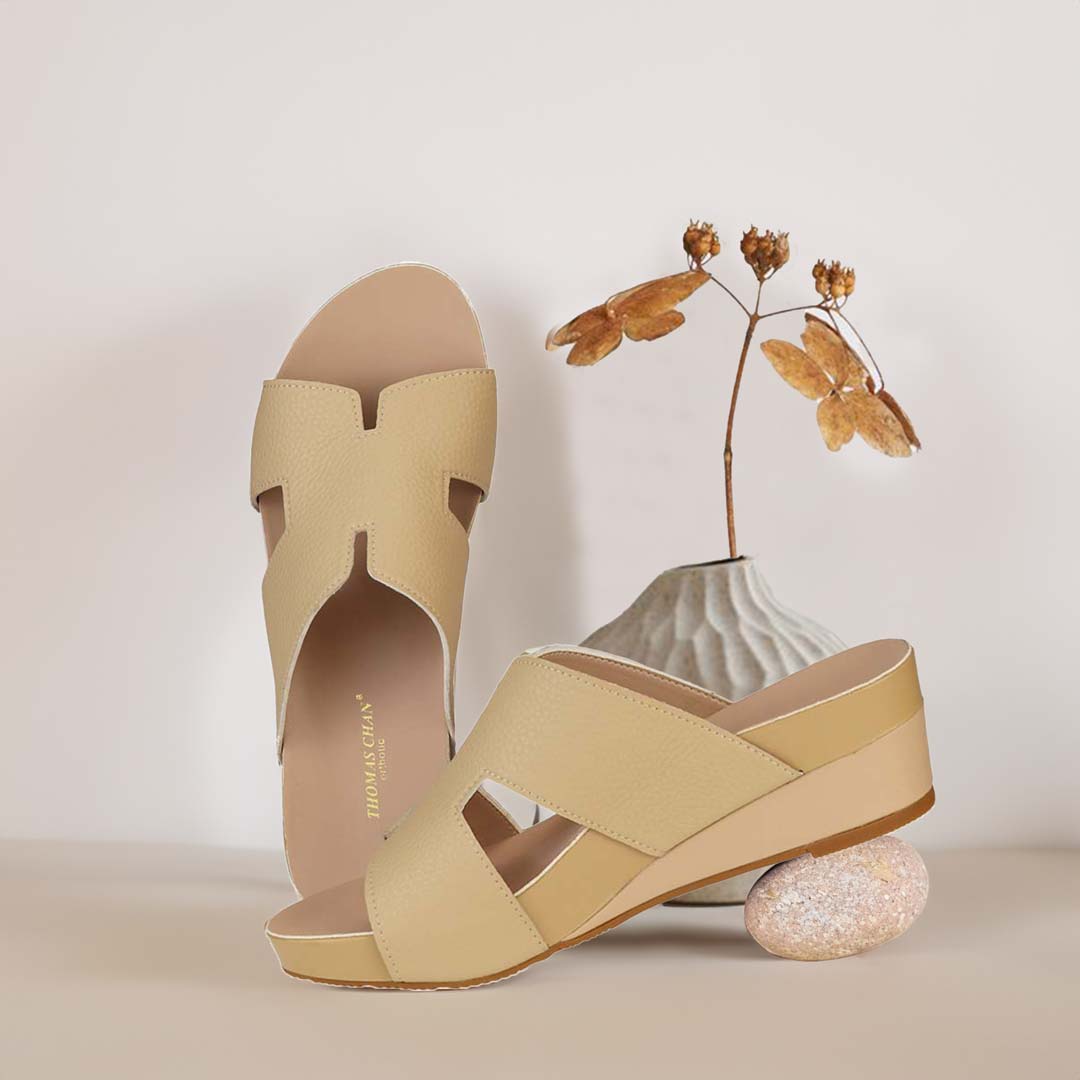 The image shows a pair of stylish Thomas Chan ivory white color comfortable Pebble Lychee H-Strap Low Wedge Slides sandals. These shoes feature a simple yet casual H-strap design, providing both elegance and comfort. The wedges are from Thomas Chan Malaysia, as indicated by the branding inside the shoe. The setting is enhanced by the presence of a few blooming flowers and a stone prop in the light background, giving the image a minimal and artistic feel