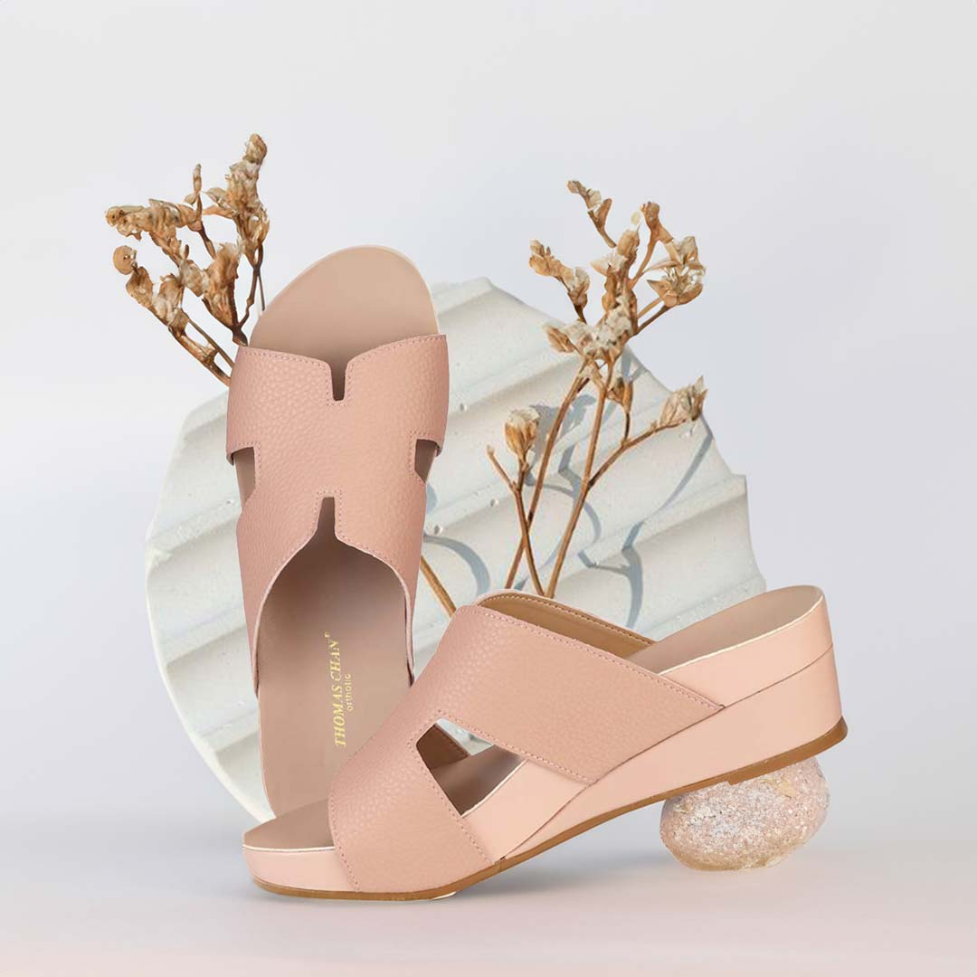 The image shows a pair of stylish Thomas Chan peachy pink color comfortable Pebble Lychee H-Strap Low Wedge Slides sandals. These shoes feature a simple yet casual H-strap design, providing both elegance and comfort. The wedges are from Thomas Chan Malaysia, as indicated by the branding inside the shoe. The setting is enhanced by the presence of a white circular grooved surface with small buds in the light background, giving the image a minimal and artistic feel.