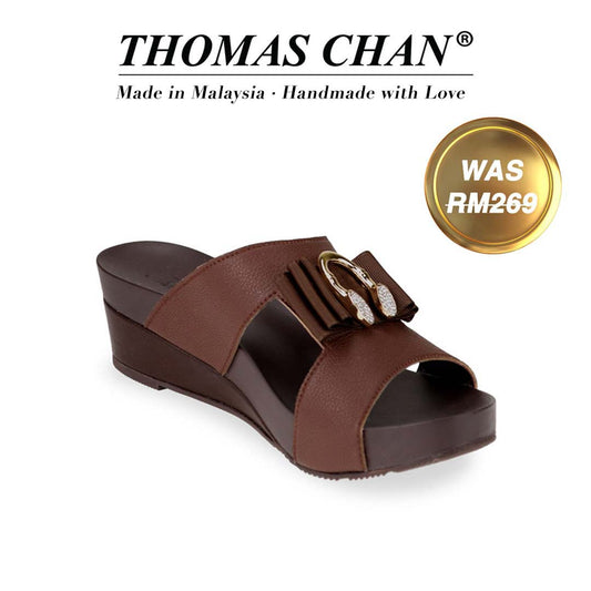  Introducing Thomas Chan's "Put-On A Smile" Sandals in brown, featuring a charming smiley buckle ribbon and a 2-inch wedge heel. Designed with built-in arch-support footbed technology for enduring comfort, these sandals are as stylish as they are supportive. A gold badge proudly displays the original price of RM 269, complemented by text celebrating "THOMAS CHAN® Made in Malaysia · Handmade with Love."